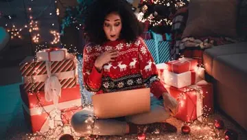 woman sitting next to Christmas tree and presents, looking at her screen